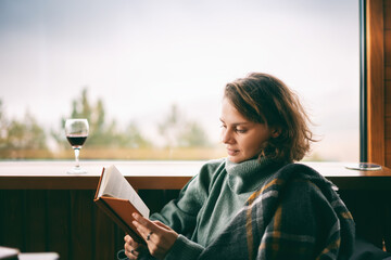 Young woman in green sweater reads a book and drinking wine near window in country house