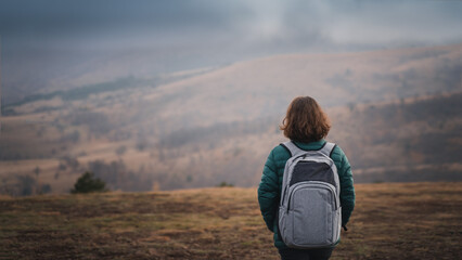 Woman traveler with gray backpack stands with her back against the background of a mountain...