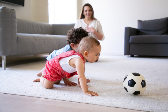 Cute little kids crawling on carpet and playing with soccer ball. Caring mother sitting on floor, smiling and watching children. Selective focus. Family indoors, weekend and childhood concept
