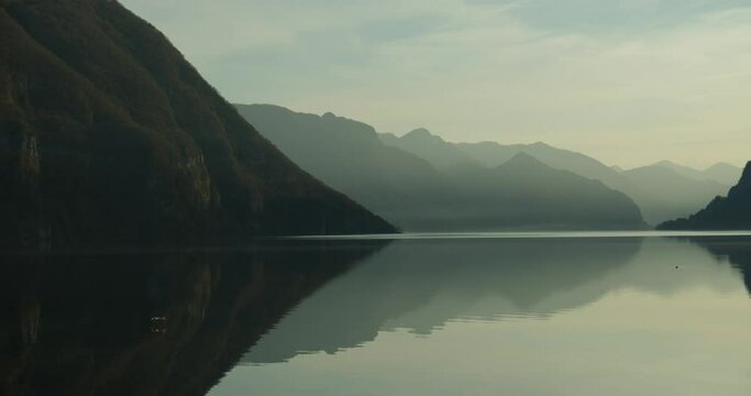 a relaxing and calming scene of a lake surrounded by mountains in northern italy; a bird sweaps in to catch a fish, but is too slow and flies away with an empty stomach. the sun is almost setting