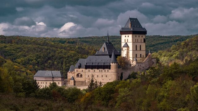 Aerial view of the beautiful Karlštejn Castle, towering solemnly on the hill surrounded by the dense green forest. Heavy gray-blue clouds rush above the gothic castle.