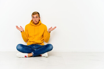 Young caucasian man sitting on the floor confused and doubtful shrugging shoulders to hold a copy space.