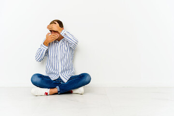 Young caucasian man sitting on the floor blink at the camera through fingers, embarrassed covering face.