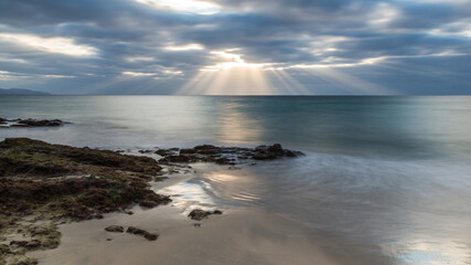 Dramatic sunrise by the sea. A seascape at sunrise with sun beams passing through coloured clouds. At foreground the sea shore with reflections of the sky in the puddles among the rocks.