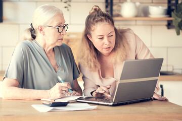 Senior woman using laptop for websurfing in her kitchen. middle-aged daughter helps her mother with documents. Mature lady sitting at work typing a notebook computer in an home office.