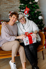 Obraz na płótnie Canvas Cheerful excited old ladies sitting on a sofa at the living room, exchanging Christmas gifts. Senior female friends having fun, laughing on Thanksgiving days. Stay active in retirement and friendship.
