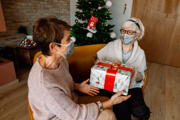 Obraz na płótnie Canvas Couple of Elderly female friends, wearing face mask, giving a Christmas present for Thanksgiving. Happy senior women with Christmas Gifts. New health regulations during Coronavirus pandemic, lockdown.