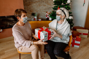 Obraz na płótnie Canvas Couple of Elderly female friends, wearing face mask, giving a Christmas present for Thanksgiving. Happy senior women with Christmas Gifts. New health regulations during Coronavirus pandemic, lockdown.