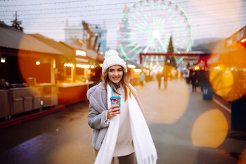 Young woman relaxing drinking coffee in  Festive Christmas fair. Smiling woman in winter style clothes walking in Christmas market. Winter holidays. Lights around.