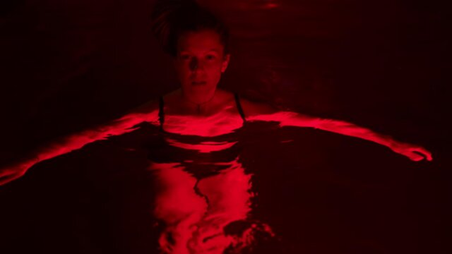 A woman floating in a dark swimming pool with red backlight seems scared. The concept of horror and suspense