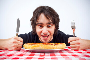 Studio photo of young man with dark hair looking down with excitement, eating a pizza, holding fork and knife.