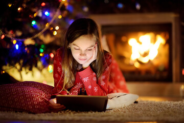 Adorable young girl using a tablet pc at home by a fireplace in warm and cozy living room on Christmas eve. Winter evening with family and kids.