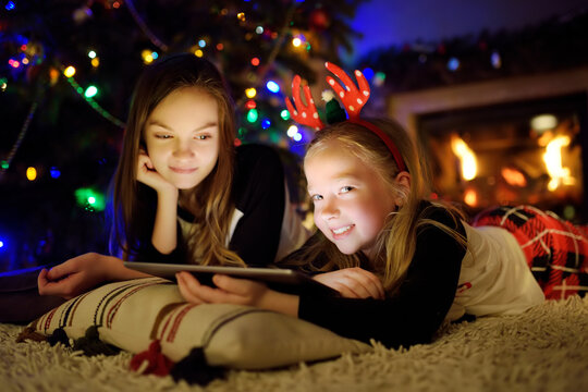 Two cute young sisters using a tablet pc at home by a fireplace in warm and cozy living room on Christmas eve. Winter evening with family and kids. - Powered by Adobe