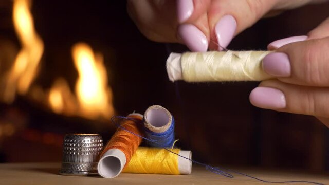 Sewing. A woman's hand takes a spool of thread, sticks a needle in it and winds the thread on top against the background of the fireplace flame. Close-up shooting.