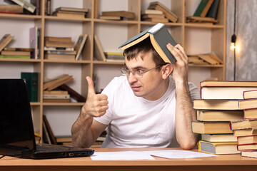 young male student sits at a table with books on the background of a cabinet and shows 