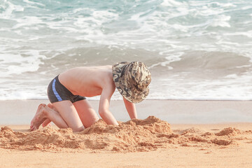 A little boy in black shorts with a blue stripe and a khaki hat on his head is digging a hole in the sand on the coast of the sea. Creating a castle. Outdoor, summer, beach, early morning.
