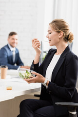  businesswoman eating meal from plastic bowl at workplace during break on blurred background