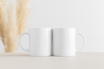 Two white mugs mockup on a beige table with pampas decoration.