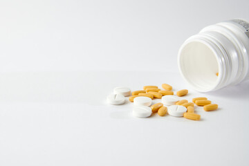 A closeup of white and yellow pills drop out of the pill bottle on the white surface
