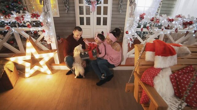 Family with little daughter and dog sitting on porch with Christmas tree and decor. Happy family concept. Young family in pullovers hugging in Christmas day. Full length shot