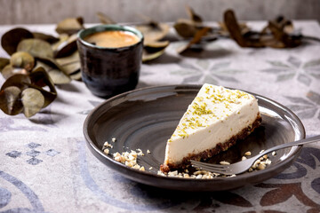 Piece of raw vegan cheesecake, no bake gluten free, decorated by lime zest and cashew nuts on plate. Cup of black coffee and Eucalyptus branches on ornate ceramic table. Sunlight