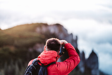 Back view of climber taking photos on top of the mountain cliff. Photographer with red jacket and backpack enjoying and looking at mountain view in Ceahlau Massif