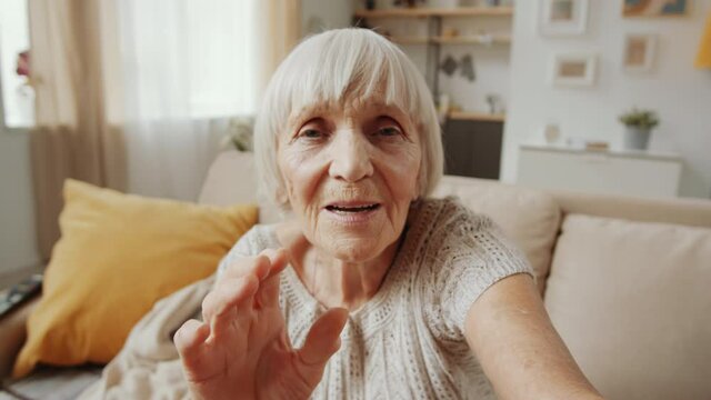 Senior grey-haired woman waving at camera and speaking on online video call while sitting on sofa in the living room