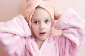 a little girl with blue eyes in a pink robe and a pink towel on her head after a bath smiles and looks into the frame. on a peach background