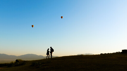 Obraz na płótnie Canvas Silhouette of two lovers on a mountainside at early morning sunrise. Balloons fly over them in the sky. A romantic painting against the backdrop of a beautiful landscape. High quality photo
