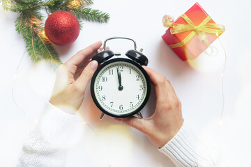 Female hands, dressed in a white sweater, hold a large black alarm clock showing twelve o`clock. On the table there is a spruce branch and Christmas tree decorations.