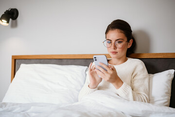 Beautiful serious girl using cellphone while sitting in bad at home