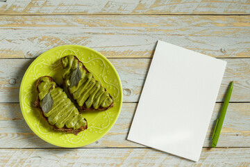 Avocado pasta with green basil, garlic and coconut cream on whole grain bread on a faience green plate next to a notebook and a fountain pen on a table of stained shabby boards