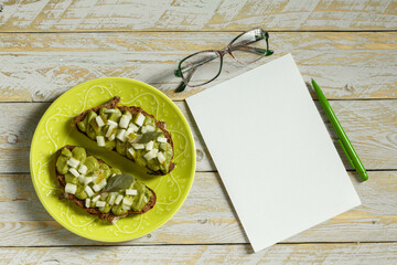 Avocado pasta with green basil, garlic and coconut cream with chopped pickled cucumbers on whole wheat bread on a green earthenware plate next to glasses, a notebook and a fountain pen on a table