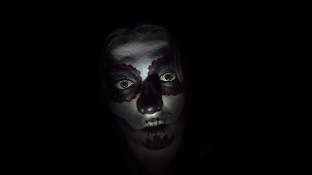Portrait of creepy woman with a make-up for Halloween in santa muerte style illuminated by red lights. A lady with painted face in close-up looks steadily and bends slowly her head on black background