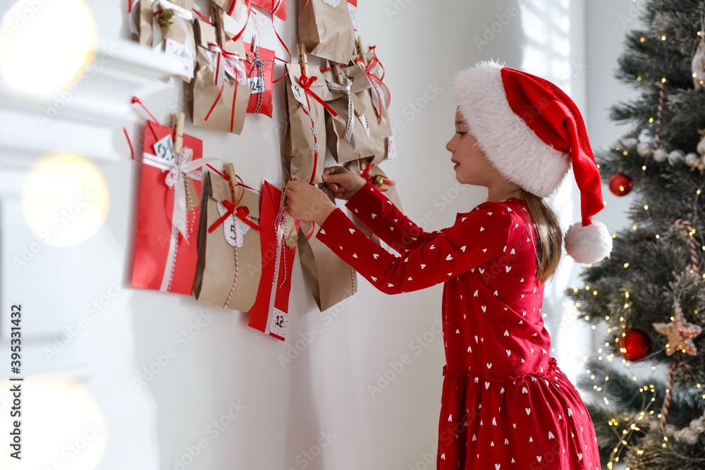 Wall mural cute little girl in santa hat taking gift from christmas advent calendar at home