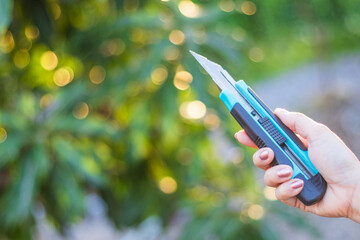 Hand holding blue cutter Knife on green natural blur background.