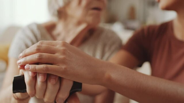 Close up selective focus shot of hands of young woman holding hands of senior grandmother while sitting with her at home and speaking