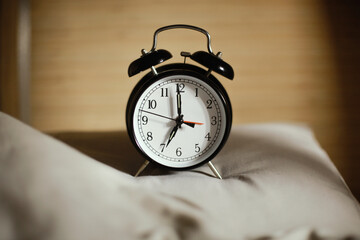 Classic black alarm clock showing seven o'clock with a bed in the background, waking up in the...