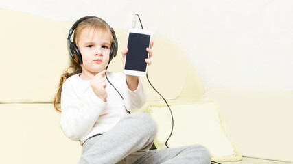 little girl sits on the sofa in the living room and listens to music with headphones, holding a smartphone in her hands. In the interior. childhood