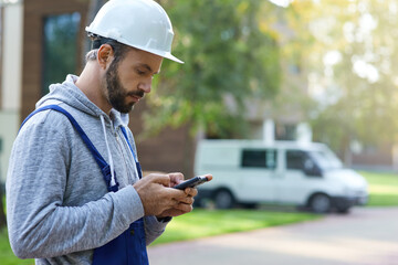 Young male builder in hardhat using smartphone, standing outdoors while working at cottage...