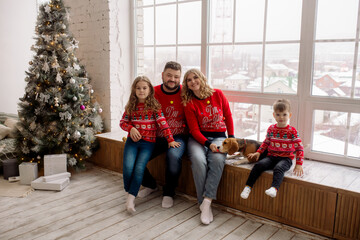 Young Caucasian beautiful family mom and dad are sitting embracing with their son, daughter and dog on the windowsill of the house among the Christmas decorations. Family portrait at home
