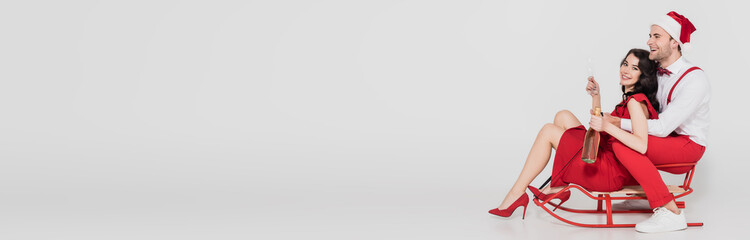  woman in dress holding bottle and glass of champagne near man in santa hat on sleigh on grey background, banner