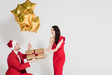 Fototapeta premium Man in santa hat holding golden balloons and gift near woman on grey background