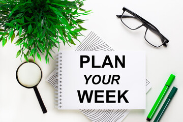 On a white background lies a notebook with the word PLAN YOUR WEEK, glasses, a magnifying glass, green markers and a green plant