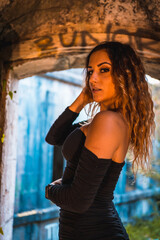 Obraz premium Lifestyle, Caucasian girl with wavy hair, in a tight black dress and heels inside a vandalized building. Portrait in window of building interior