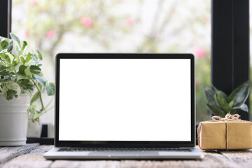 laptop mock up white blank screen with  gift box and plant pot on wooden table in front of windows 
