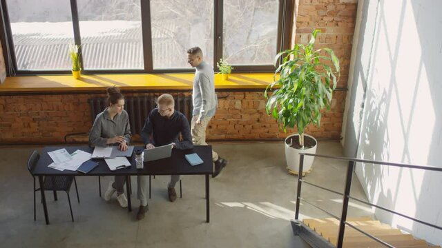 Top view footage of young specialists working on laptop at desk by big window in office loft and supervisor helping them while going by