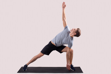 Strong young man standing in yoga pose on sports mat over light studio background, full length portrait