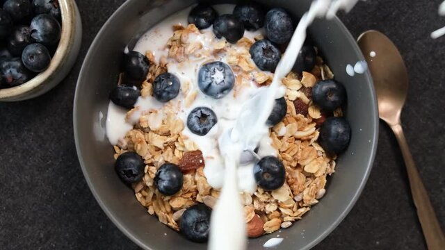 Slow motion of milk pouring in bowl of crunchy oat granola. Healthy breakfast cereals with blueberries and milk
