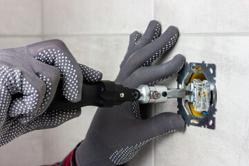 Gray-gloved hands hold a screwdriver and screw the contacts into an electrical outlet.